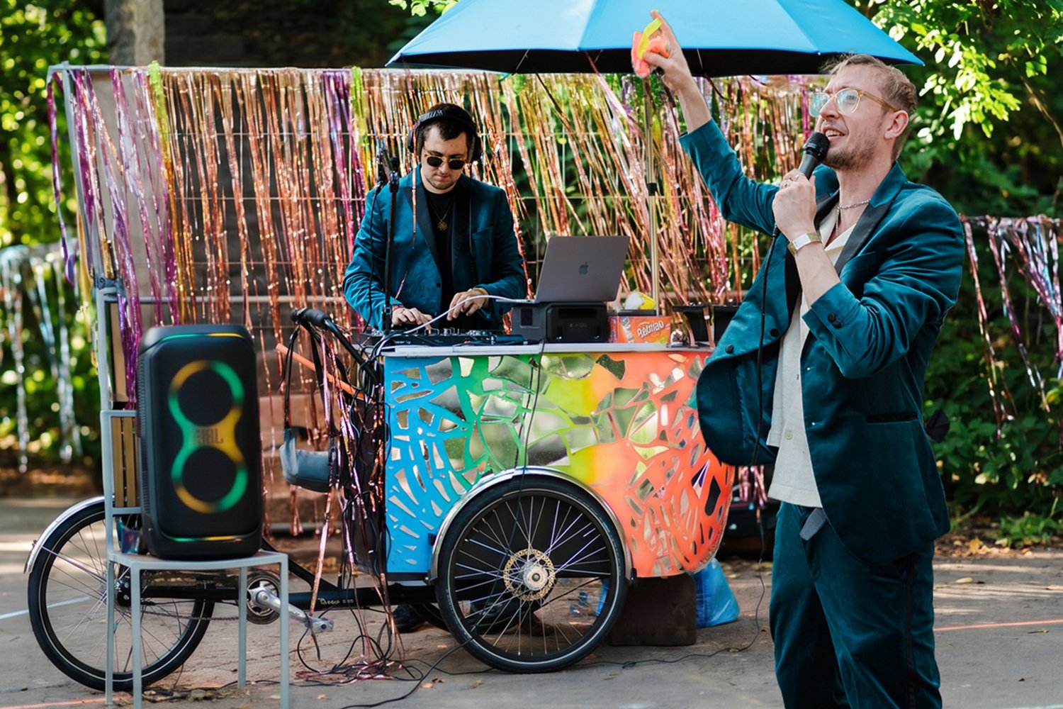 Zwei Personen an einer mobilen Discoshow-Station im Freien. Ein DJ steht hinter einem bunten Mischpult, eine weitere Person spricht mit Mikrofon und erhobener Hand davor. Ein großer Lautsprecher steht links.