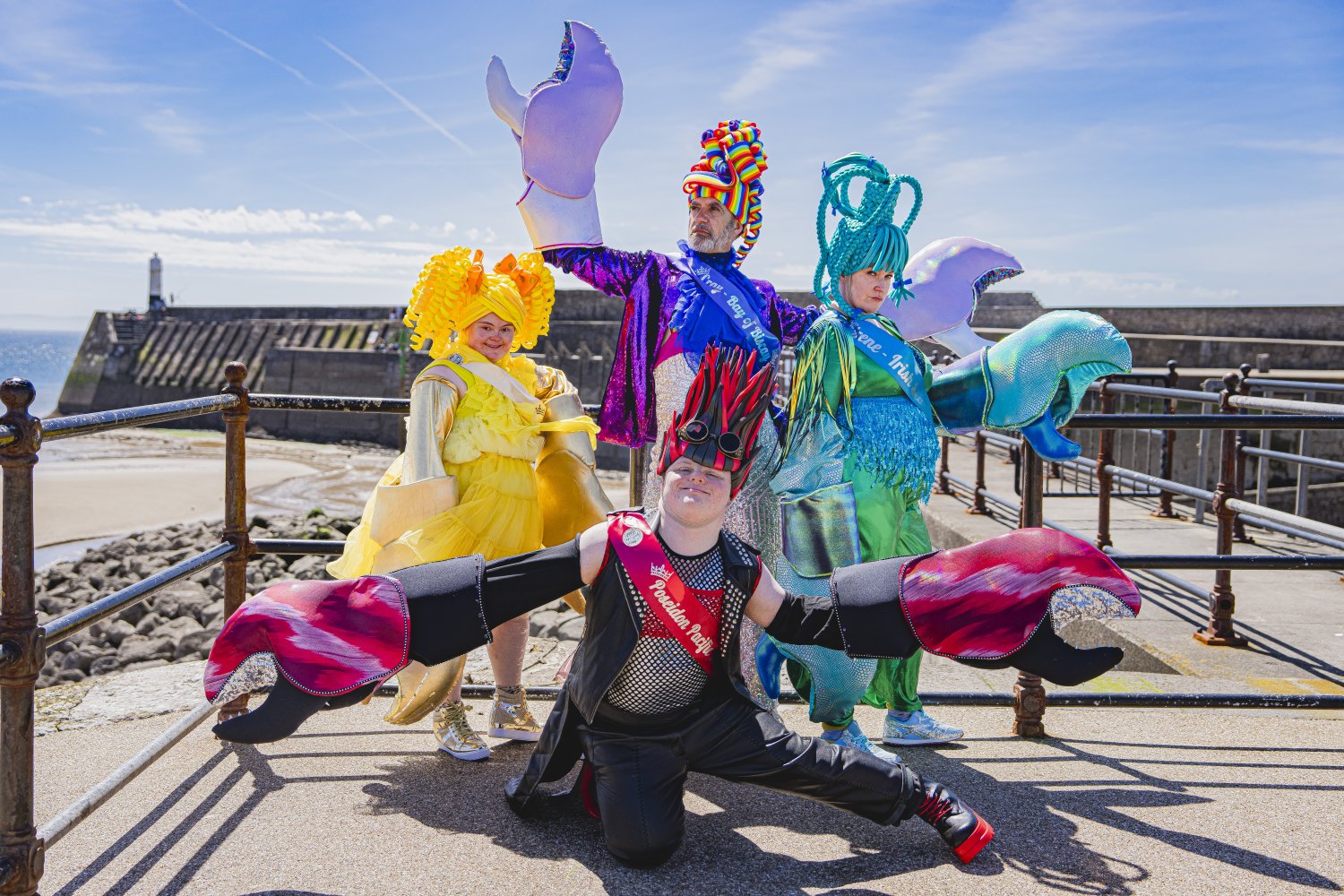 Vier Personen in extravaganten, bunten Krabbenkostümen posieren bei Sonnenschein auf einer Strandpromenade. Vorne kniet eine Person in einem schwarz-roten Punk-Outfit mit großen, gepolsterten Krabbenscheren. Dahinter stehen drei Personen in auffälligen ge