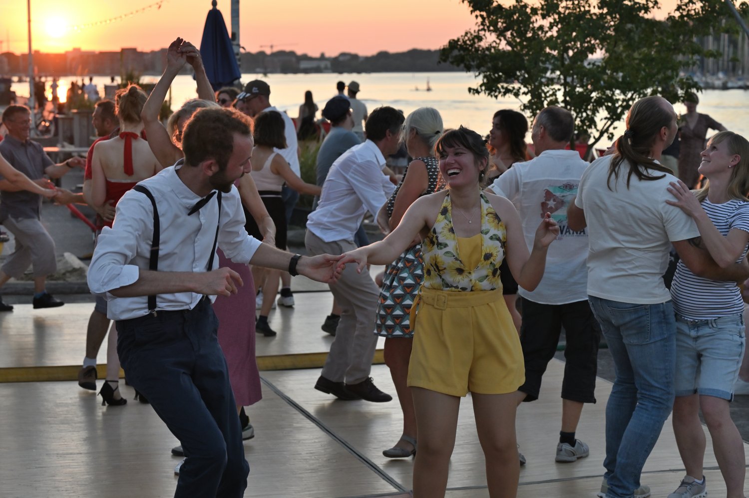 Ein lachendes Paar tanzt Swing auf einem Holzdeck am Wasser während des Sonnenuntergangs. Die vordere Person trägt ein gelbes Outfit mit Blumenmuster, die hintere ein weißes Hemd mit Fliege und Hosenträgern. Viele andere Tanzpaare sind im Hintergrund zu s