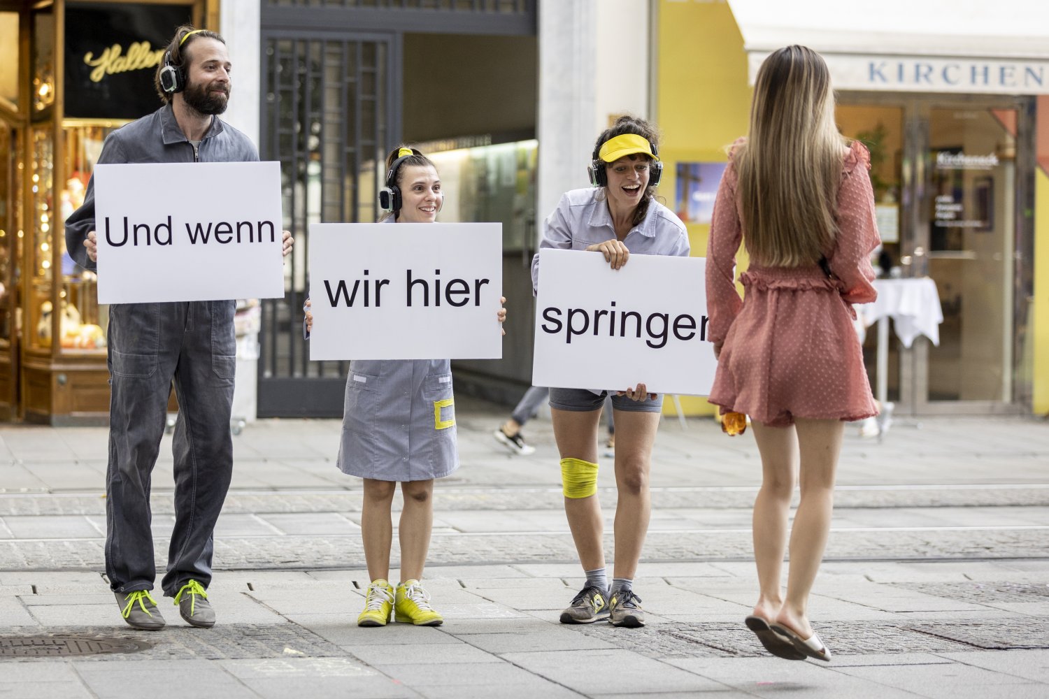 Drei Personen in Arbeitskleidung und Kopfhörern stehen in einer Fußgängerzone und halten Schilder mit dem Text "Und wenn wir hier springen". Eine vierte Person in einem rosa Kleid springt rechts vorbei.