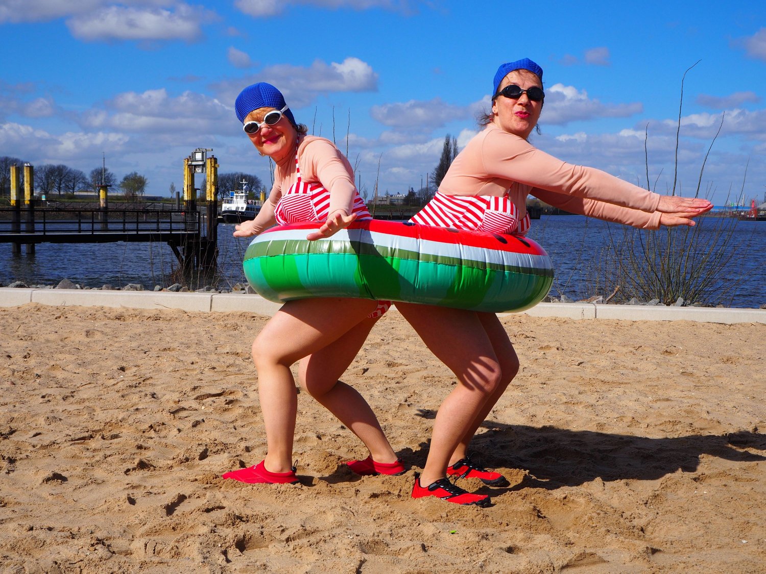Zwei Personen in rot-weiß gestreifter Badekleidung und blauen Badekappen stehen an einem Sandstrand Rücken an Rücken. Sie stecken gemeinsam in einem großen Schwimmreifen im Wassermelonen-Design und haben die Arme zur Seite ausgestreckt.