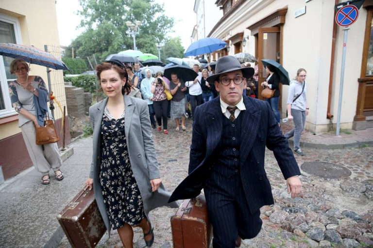 Zwei Personen in Vintage-Kleidung gehen mit alten Koffern auf einem Kopfsteinpflasterweg. Im Hintergrund eine Menschenmenge mit Regenschirmen.