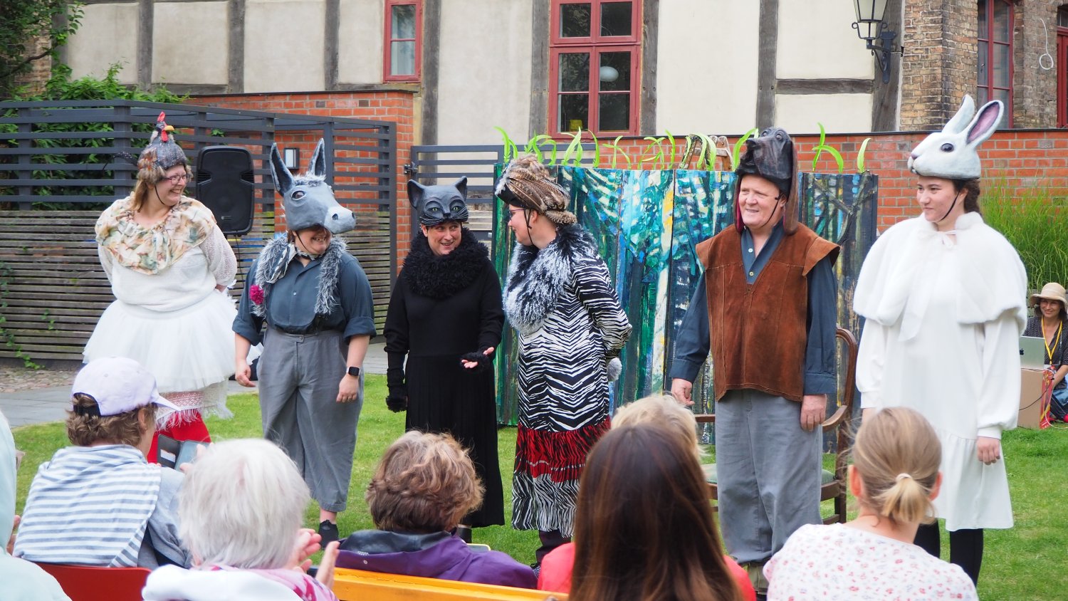 Eine Theateraufführung im Freien. Sechs Personen in unterschiedlichen Kostümen und mit Tiermasken oder Tierkopfbedeckungen stehen nebeneinander auf einer Rasenfläche. Im Vordergrund sitzen Zuschauer*innen und verfolgen das Stück. Im Hintergrund stehen ein