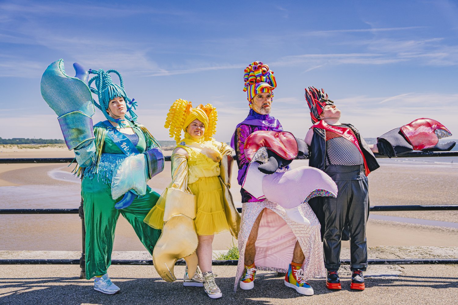 Vier Personen in farbenfrohen Krabbenkostümen stehen nebeneinander an einem Geländer, im Hintergrund ist ein weitläufiger Sandstrand zu sehen. Sie tragen detailreiche Outfits in Grün-Blau, Gelb, Lila mit Regenbogen-Kopfschmuck sowie Schwarz-Rot und streck