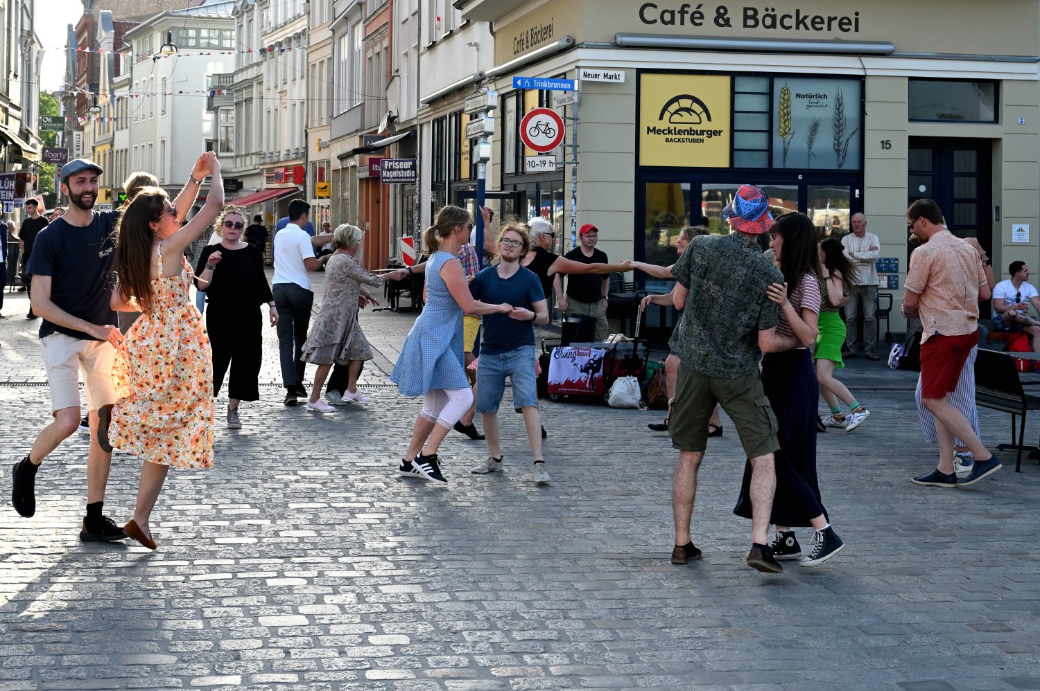 Menschen tanzen Swing auf einem gepflasterten Platz in einer europäischen Stadt. Ein Café und Bäckerladen ist im Hintergrund. Mehrere Tanzpaare und Zuschauer*innen sind zu sehen. Die Sonne scheint.