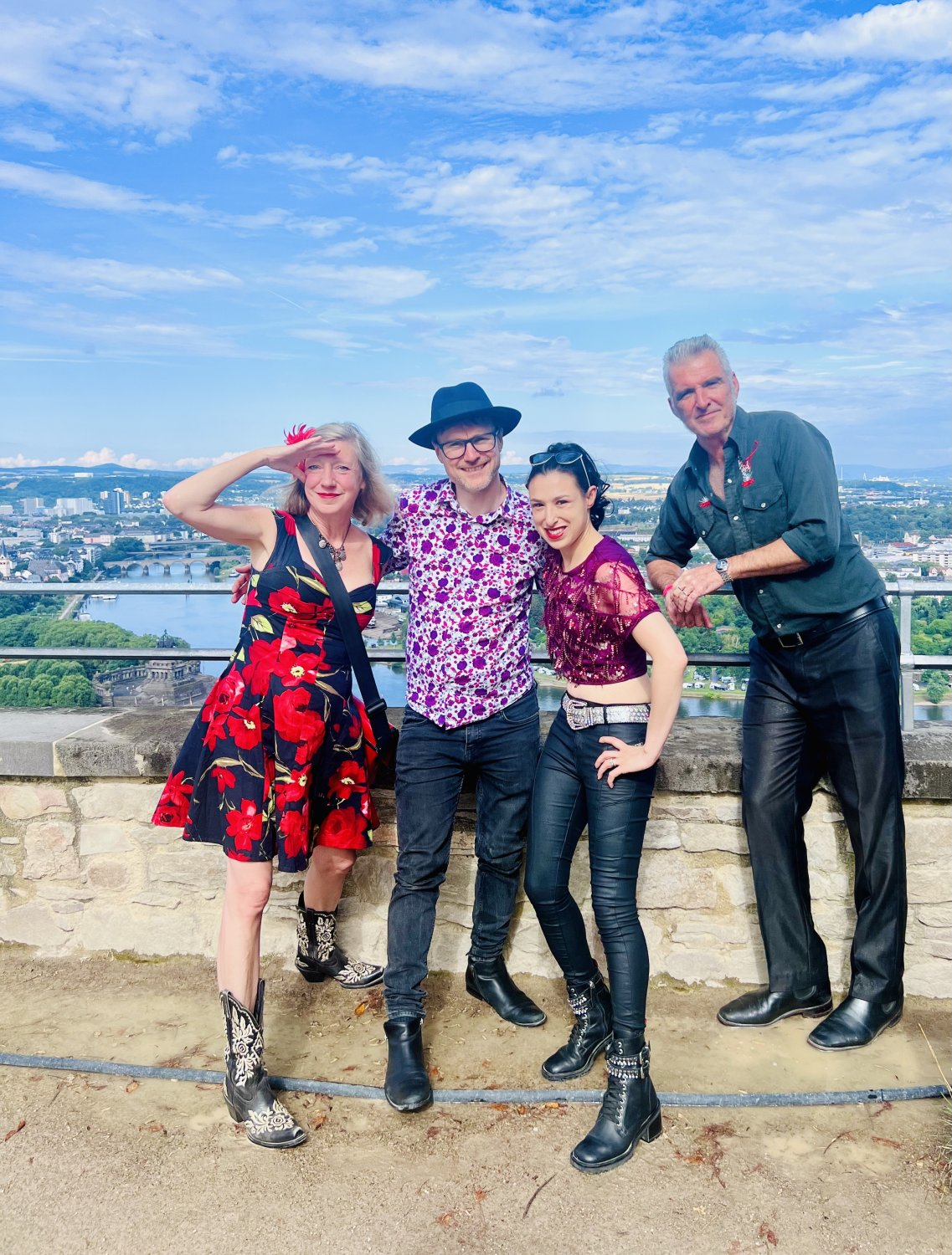 Vier Personen einer Musikgruppe stehen auf einer Aussichtsplattform über einer Stadt und einem Fluss. Sie tragen bunte Kleidung und posieren lächelnd für das Foto unter blauem Himmel.