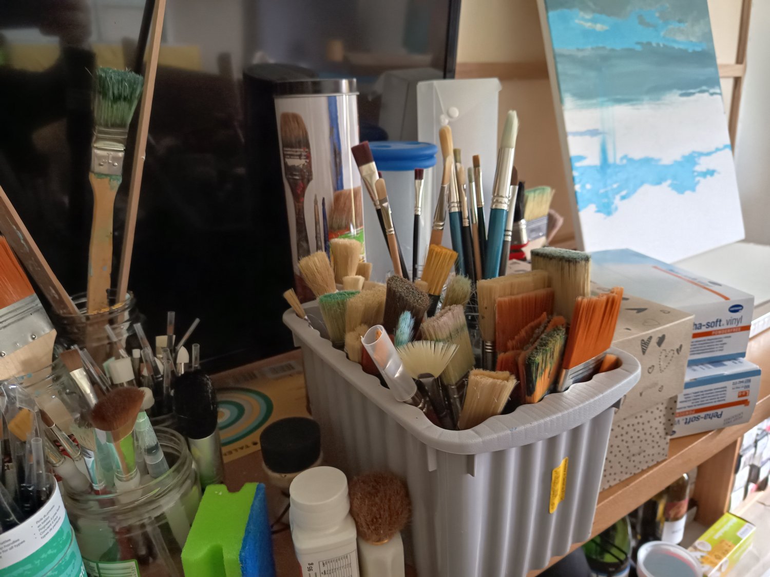 Nahaufnahme eines Arbeitsplatzes für Künstler*innen. In einer grauen Box und in Gläsern stecken dicht an dicht zahlreiche Pinsel in unterschiedlichen Größen und Formen. Auf dem Tisch liegen zudem Farbdosen, ein Schwamm und im Hintergrund steht eine Leinwa