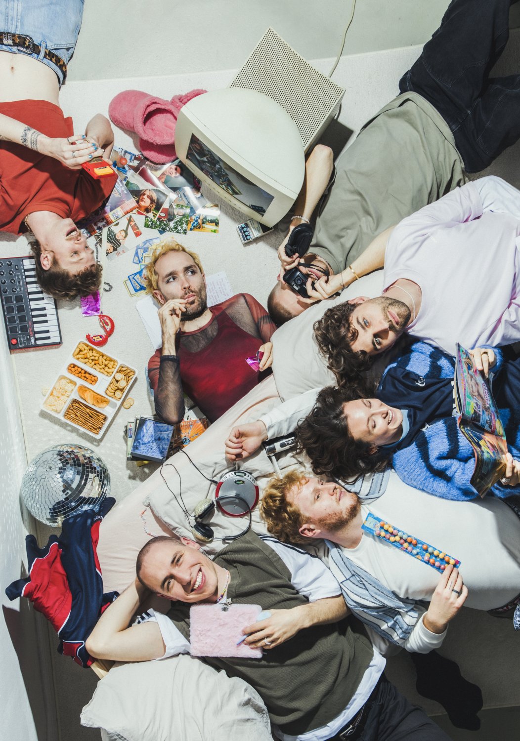 Acht junge Personen liegen in einer Gruppe auf einem Bett oder dem Boden, umgeben von Objekten wie einem Röhrenfernseher, Snackplatten, einer Discokugel und Magazinen. Sie blicken direkt in die Kamera, teilweise lächelnd. Eine Person hält ein rosa Plüschb