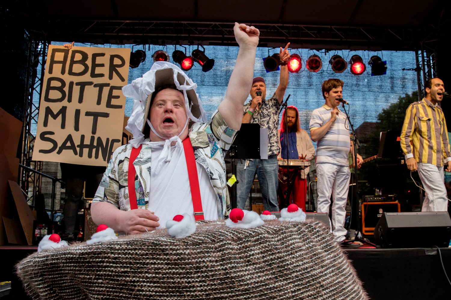 Live-Konzertfoto einer Person im Vordergrund mit weißer Haube und roten Hosenträgern, die triumphierend singt. Im Hintergrund steht eine Schlagzeugerin mit einem Schild "ABER BITTE MIT SAHNE". Die Bühne ist rot beleuchtet.