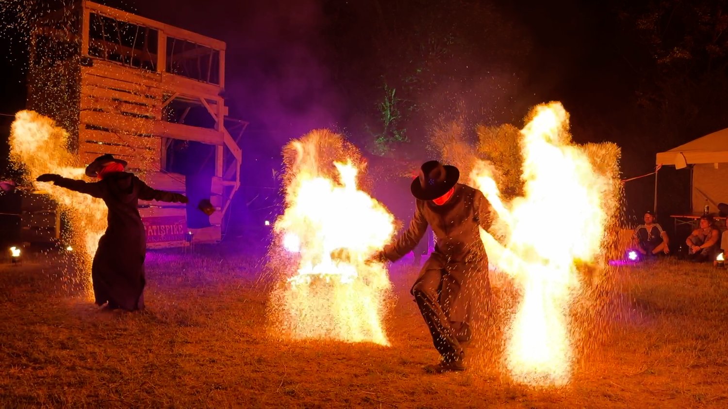 Eine nächtliche Feuershow mit zwei Personen in Mänteln und Hüten, die in großen Flammenwolken und sprühenden Funken stehen. Im Hintergrund sind eine Holzstruktur, ein Zelt und sitzende Zuschauer*innen zu sehen.