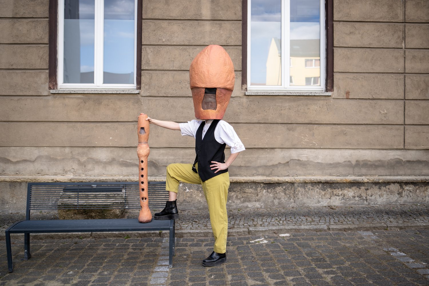 Eine Person posiert auf einem Kopfsteinpflaster vor einer Hausfassade mit zwei Fenstern. Sie trägt eine große, orangefarbene Blockflöten-Mundstück-Maske, ein weißes Hemd, eine schwarze Weste und eine gelbe Hose. Die Person hat einen Fuß auf eine dunkle Me